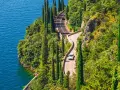 Kręta droga z samochodami wzdłuż skalistego wybrzeża jeziora Garda, otoczona cyprysami i bujną zielenią.