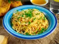 Pyszne spaghetti aglio olio posypane parmezanem i natką pietruszki, podane w niebieskiej misce. Obok grzanki czosnkowe i kawałek sera.