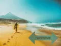 Kobieta w bikini idzie po piaszczystej plaży Fuerteventury z butami w ręku, w tle góry i ocean.
