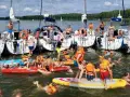Ludzie w kamizelkach ratunkowych i strojach kąpielowych pływają i bawią się na wodzie przy żaglówkach.