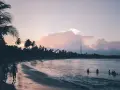 Zachód słońca na Sri Lance we wrześniu. Surferzy idą plażą, w oceanie kąpią się ludzie. Palmy i chmury w pastelowych barwach.