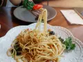 Widok z bliska na talerz z makaronem aglio olio, z widelcem unoszącym porcję spaghetti z bazylią i pomidorkiem.