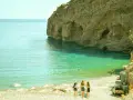 Trzy dziewczyny idące po kamienistej plaży w kierunku turkusowego morza przy skalistym klifie.