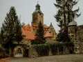 Kamienny zamek z wieżą i czerwonymi dachami, otoczony drzewami i murem.