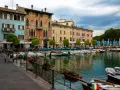 Kolorowe kamienice nad jeziorem w Desenzano del Garda, z łodziami zacumowanymi przy brzegu i stolikami restauracyjnymi na promenadzie.