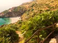 Widok na zatoczkę z piaszczystą plażą, turkusową wodą i domkiem na wzgórzu, otoczone bujną roślinnością.
