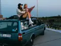 Dwie kobiety siedzą na dachu niebieskiego kombi przy nabrzeżu, jedna wskazuje palcem w dal.