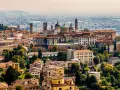 Widok na **bergamo stare miasto** z wieżami i kopułami, otoczone zielenią i innymi budynkami.