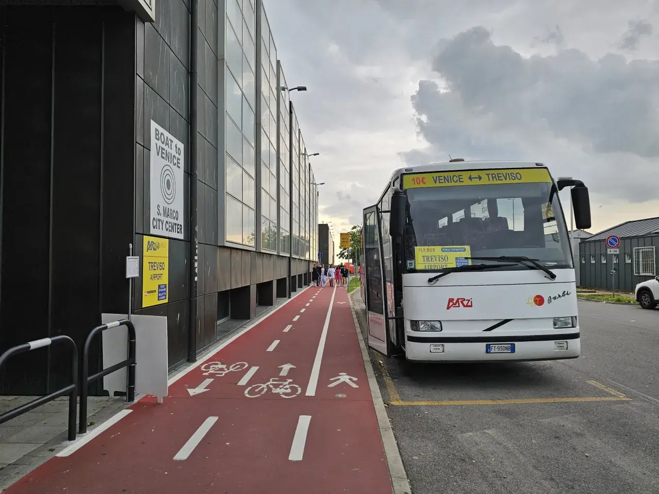 Autobus z napisem "Venice Treviso" czeka na pasażerów przy lotnisku. Obok czerwona ścieżka rowerowa i piesza.
