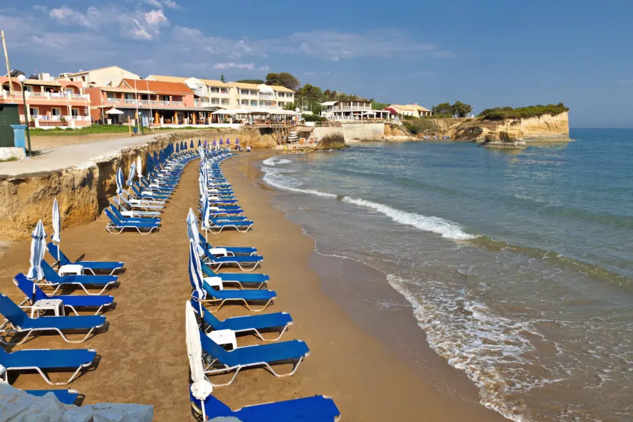 Rzędy leżaków i parasoli na piaszczystej plaży, z hotelami w tle. Idealne miejsce, by odpocząć i zastanowić się, gdzie na Korfu wybrać się na wakacje.