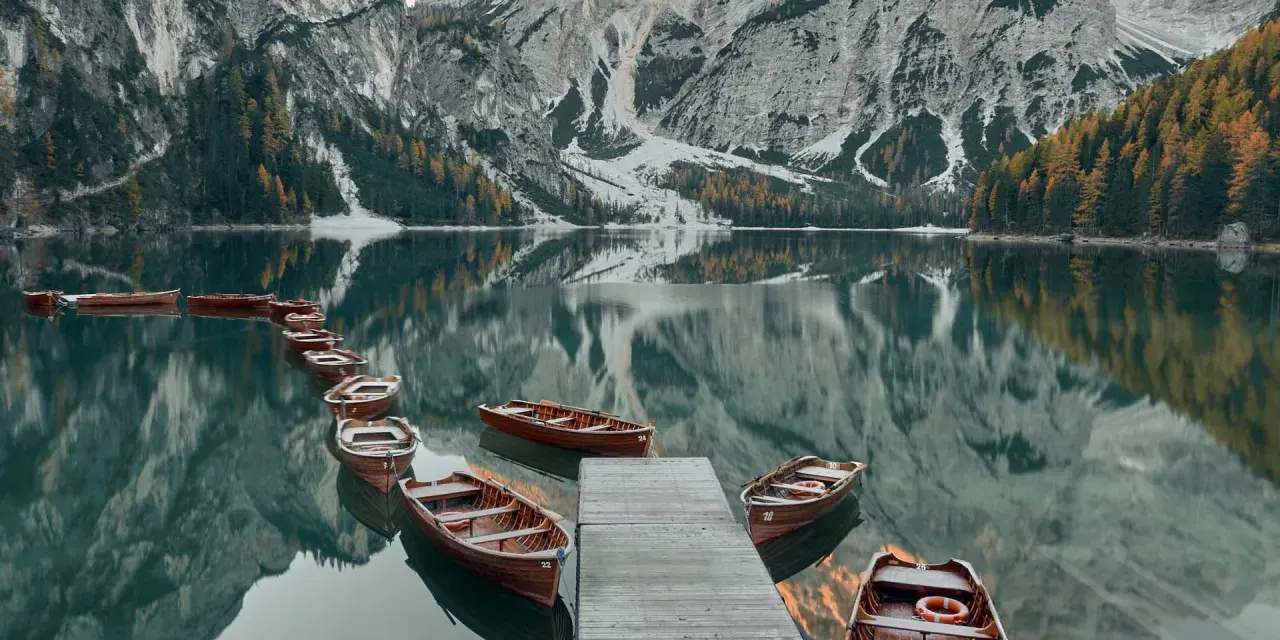 Drewniane łodzie przy drewnianym pomoście na spokojnym Lago di Braies, odbijające się w tafli wody, z majestatycznymi górami w tle.