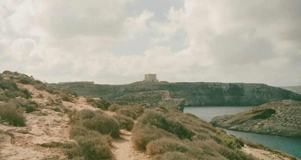 Malta czy warto? Kamienista ścieżka prowadzi przez skaliste wybrzeże z niską roślinnością do zatoki. W oddali widać wieżę obronną.