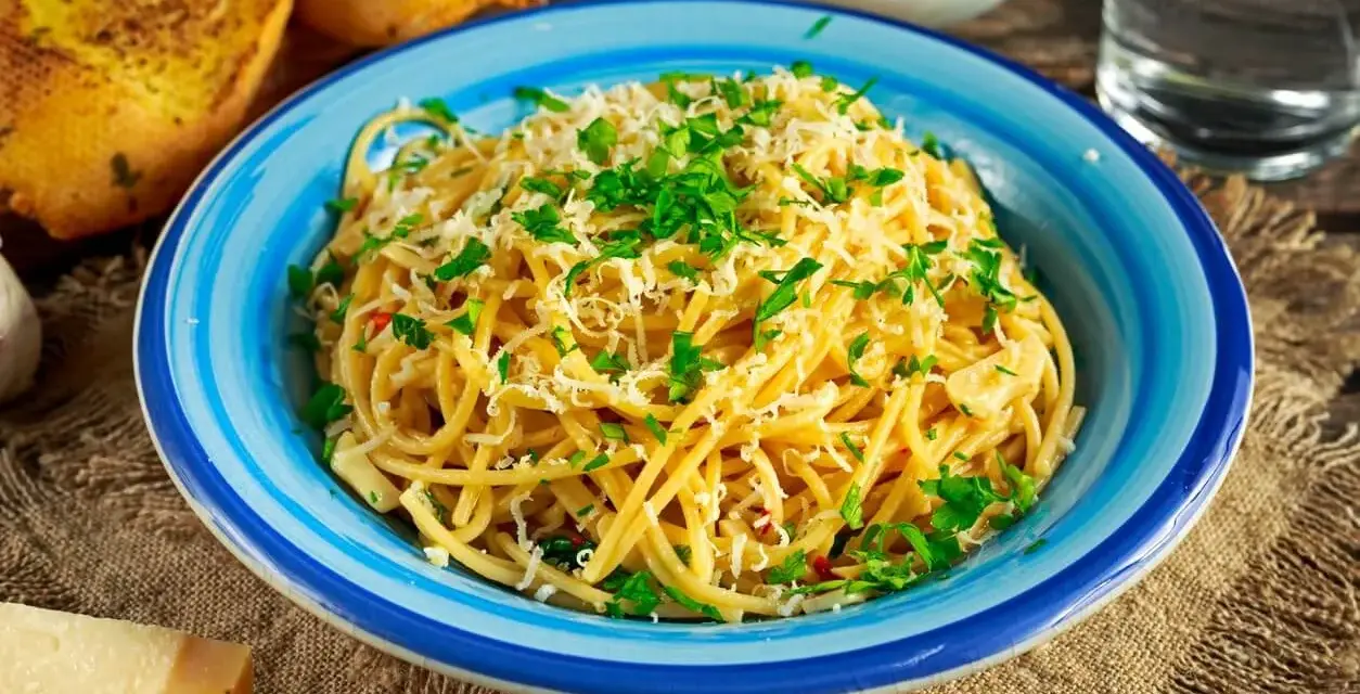 Pyszne spaghetti aglio olio posypane parmezanem i natką pietruszki, podane w niebieskiej misce. Obok grzanki czosnkowe i kawałek sera.