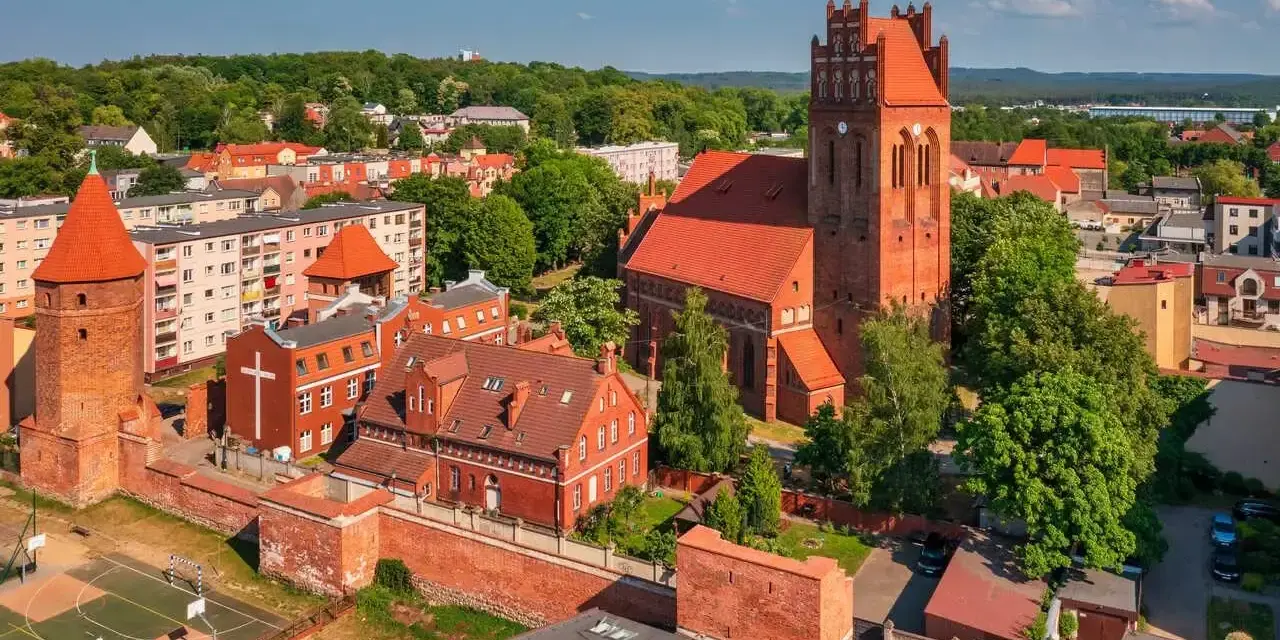 Zamek krzyżacki w Lęborku, z charakterystyczną wieżą i ceglaną architekturą, otoczony zielenią i zabudowaniami miejskimi.