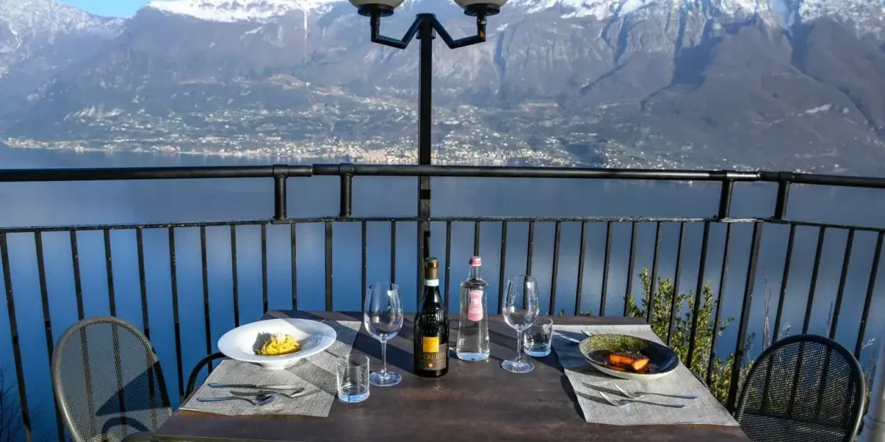 Stół z winem i jedzeniem na tarasie z widokiem na jezioro i ośnieżone góry. Idealne miejsce na romantyczną kolację z widokiem, prawdziwa "terrazza del brivido".