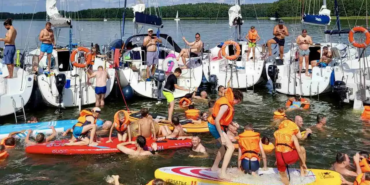 Ludzie w kamizelkach ratunkowych i strojach kąpielowych pływają i bawią się na wodzie przy żaglówkach.