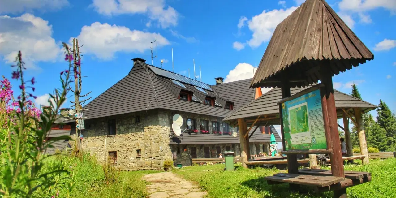 Kamienny schronisko na Turbaczu z dzieckiem w tle, otoczone zielenią i kwitnącymi kwiatami.