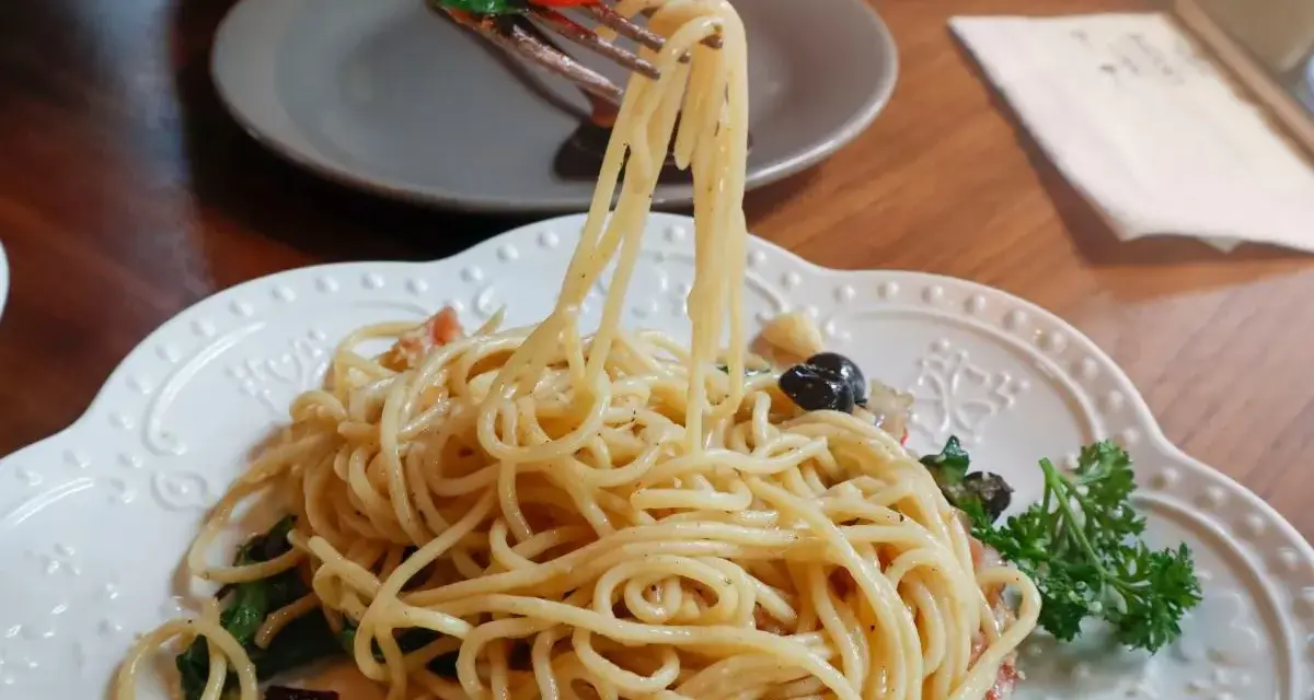 Widok z bliska na talerz z makaronem aglio olio, z widelcem unoszącym porcję spaghetti z bazylią i pomidorkiem.