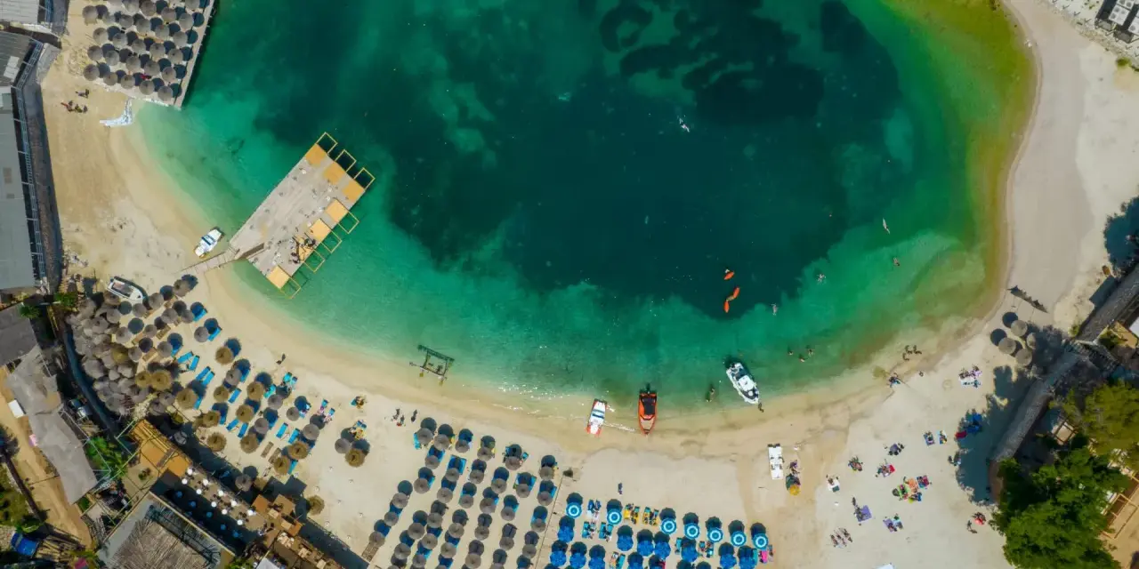 Widok z lotu ptaka na zatokę z turkusową wodą, plażą z leżakami i parasolami oraz łodziami.