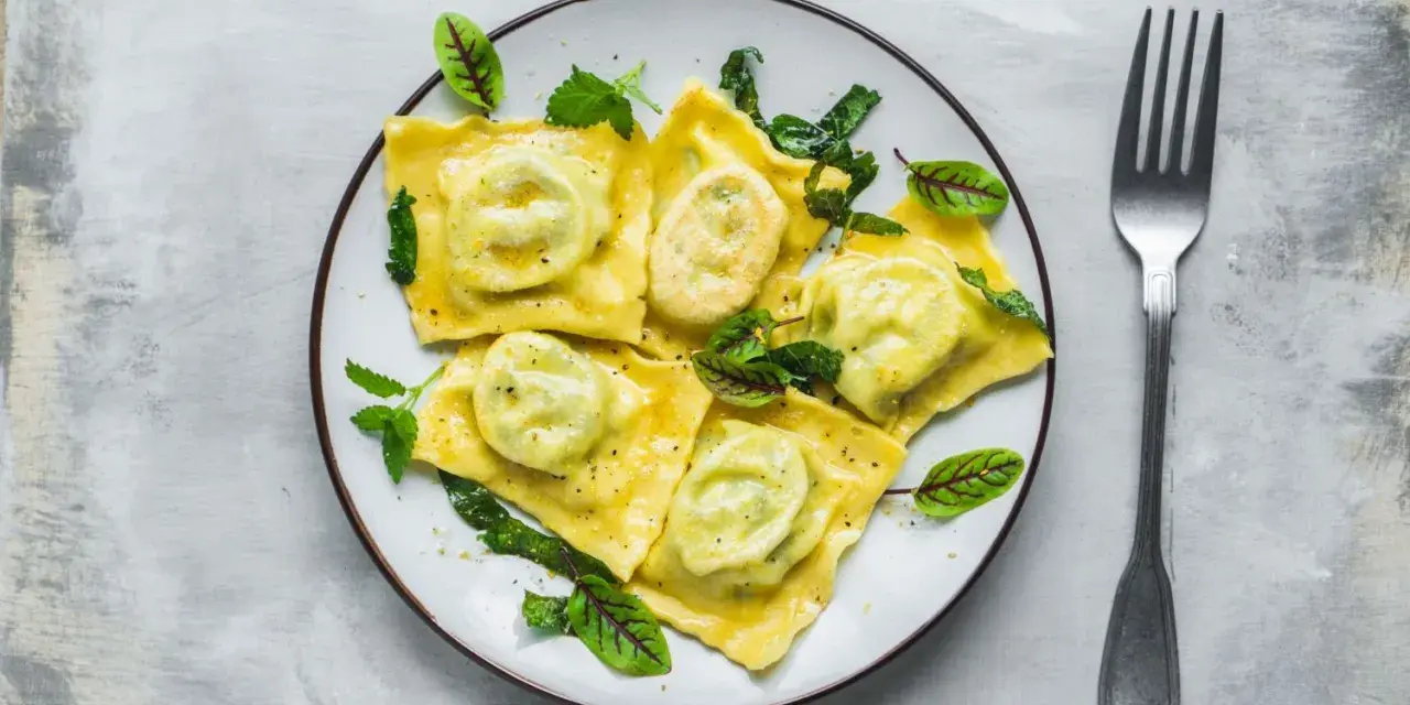 Pyszne ravioli z ricottą i szpinakiem, polane sosem, ozdobione świeżymi ziołami. Obok leży widelec.