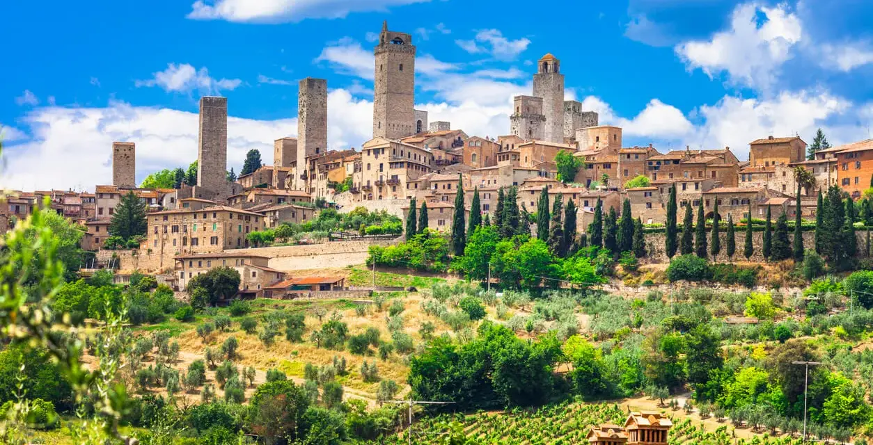 Widok na średniowieczne wieże i zabudowania San Gimignano w Toskanii, otoczone zielenią i winnicami.