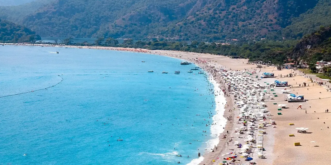 Plaża z turkusową wodą, górami w tle i tłumem ludzi odpoczywających pod parasolami.