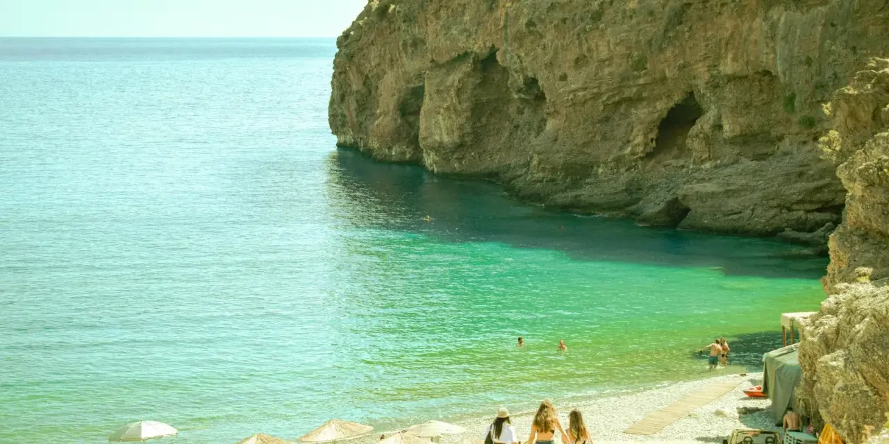 Trzy dziewczyny idące po kamienistej plaży w kierunku turkusowego morza przy skalistym klifie.