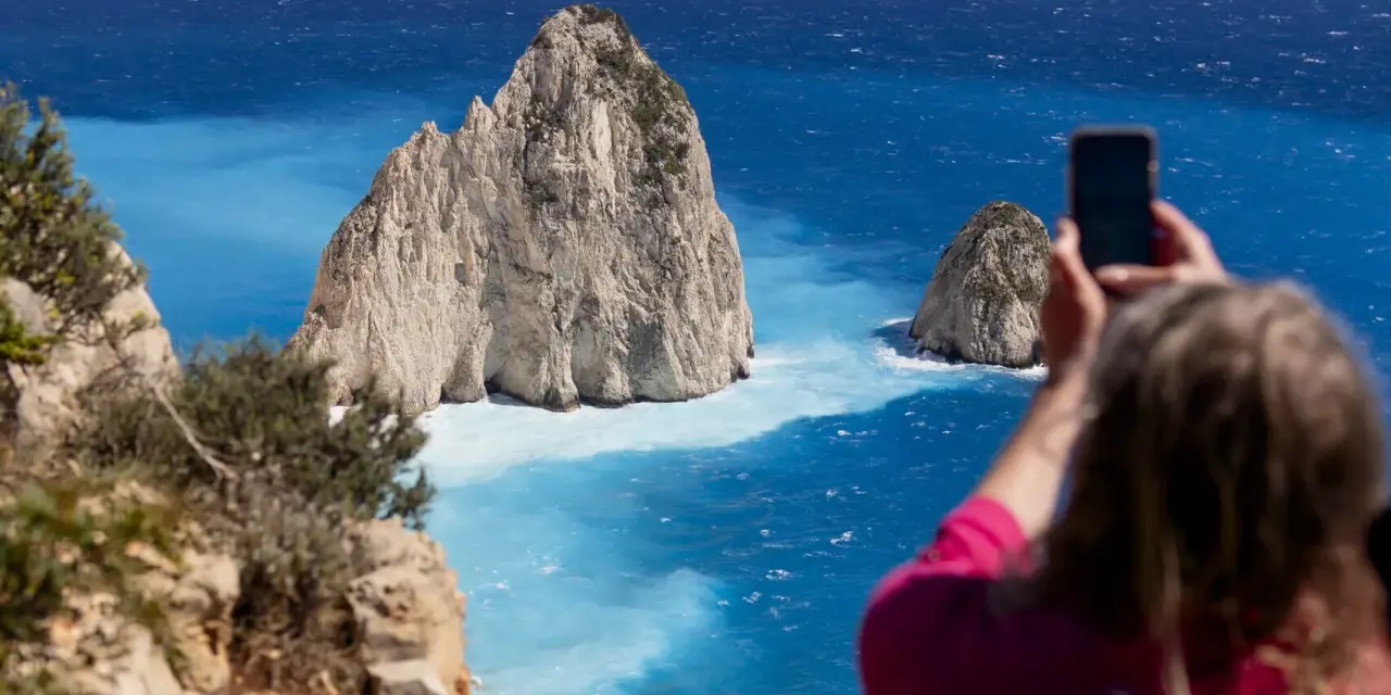 Widok na turkusowe morze i skaliste wysepki. Kobieta robi zdjęcie telefonem. Zakynthos czy warto? Tak, warto!