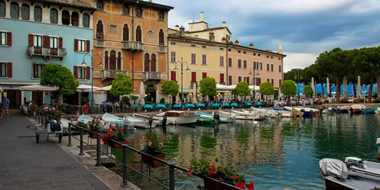 Kolorowe kamienice nad jeziorem w Desenzano del Garda, z łodziami zacumowanymi przy brzegu i stolikami restauracyjnymi na promenadzie.