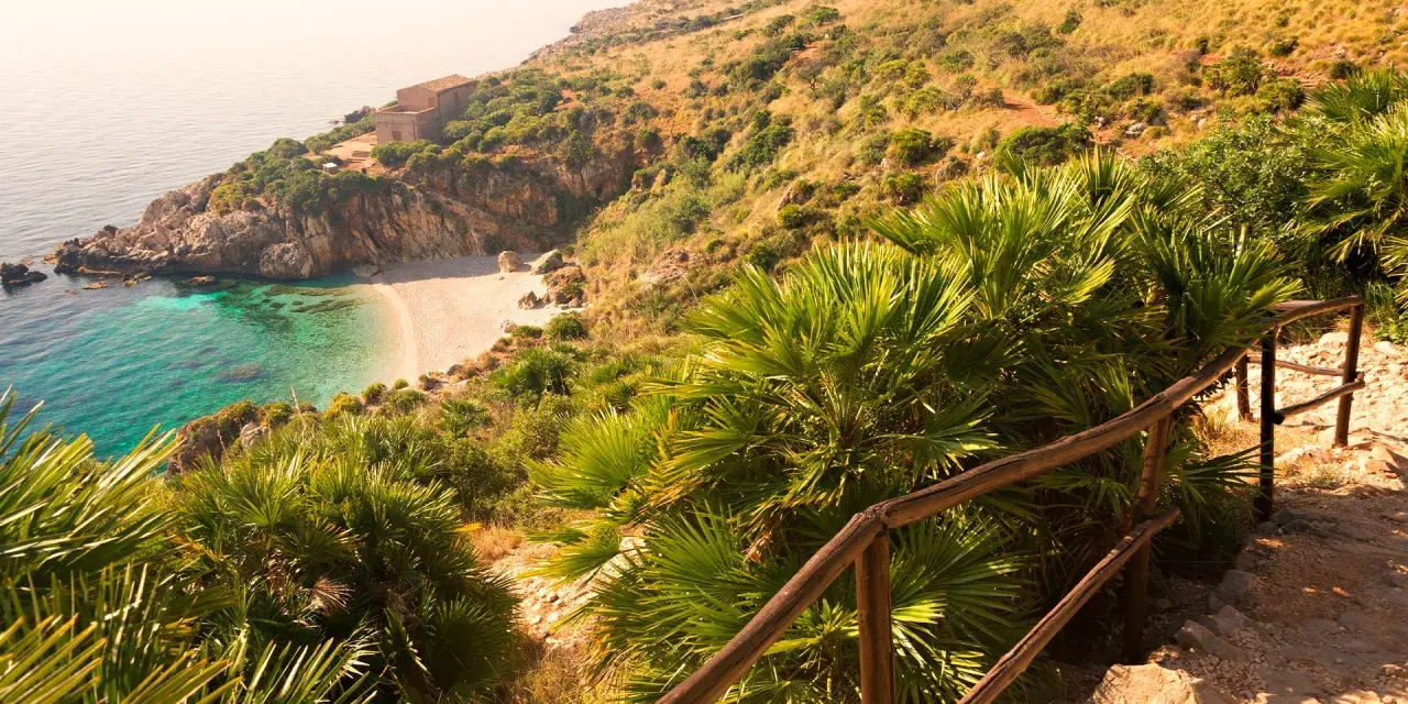 Widok na zatoczkę z piaszczystą plażą, turkusową wodą i domkiem na wzgórzu, otoczone bujną roślinnością.