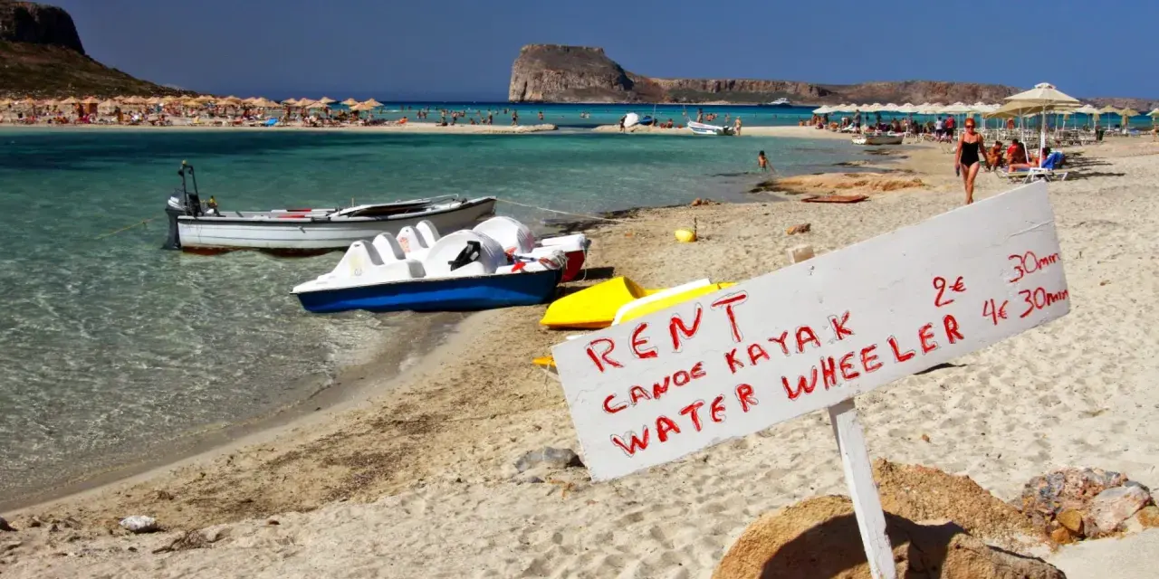Maj na Krecie: plaża z leżakami, turkusowa woda, łódki i tablica z ofertą wynajmu kajaków i rowerów wodnych.