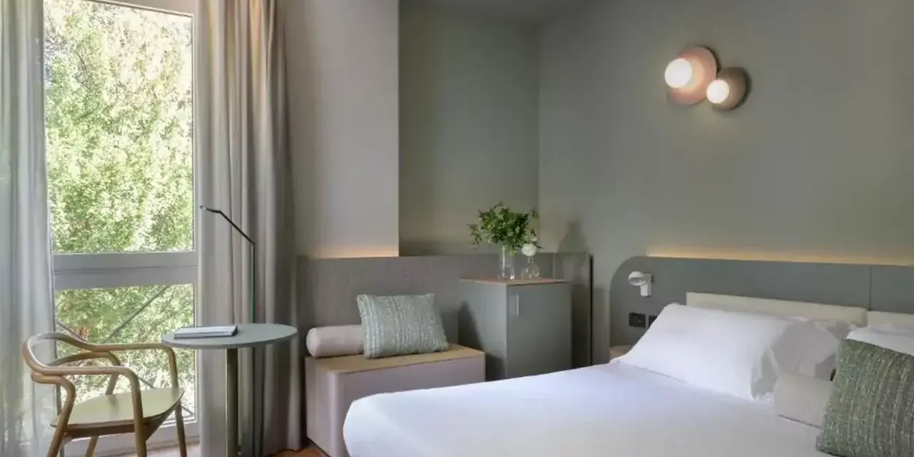 Nowoczesny pokój hotelowy z łóżkiem, fotelem i oknem z widokiem na zieleń.