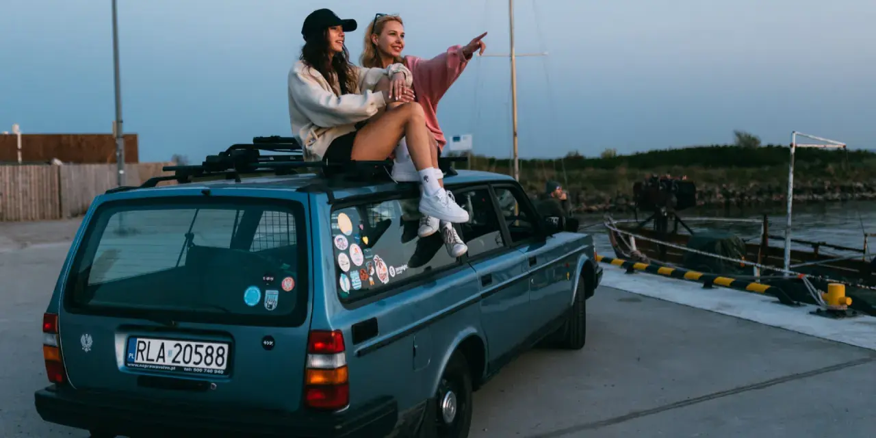 Dwie kobiety siedzą na dachu niebieskiego kombi przy nabrzeżu, jedna wskazuje palcem w dal.