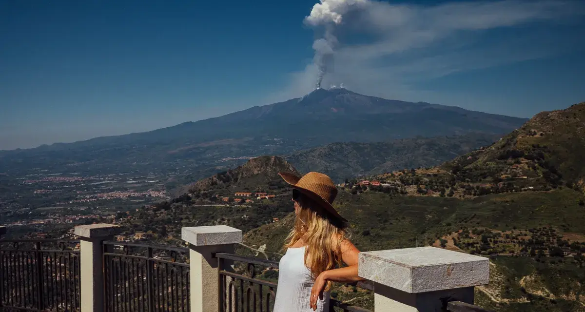 Kobieta w kapeluszu patrzy na dymiący wulkan Etna z tarasu widokowego.