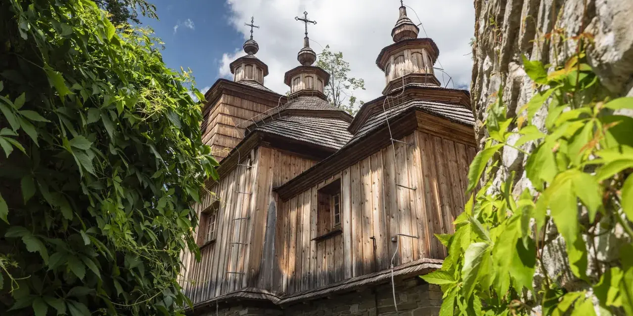 Drewniana cerkiew z trzema kopułami, otoczona zielenią, na tle błękitnego nieba z chmurami.