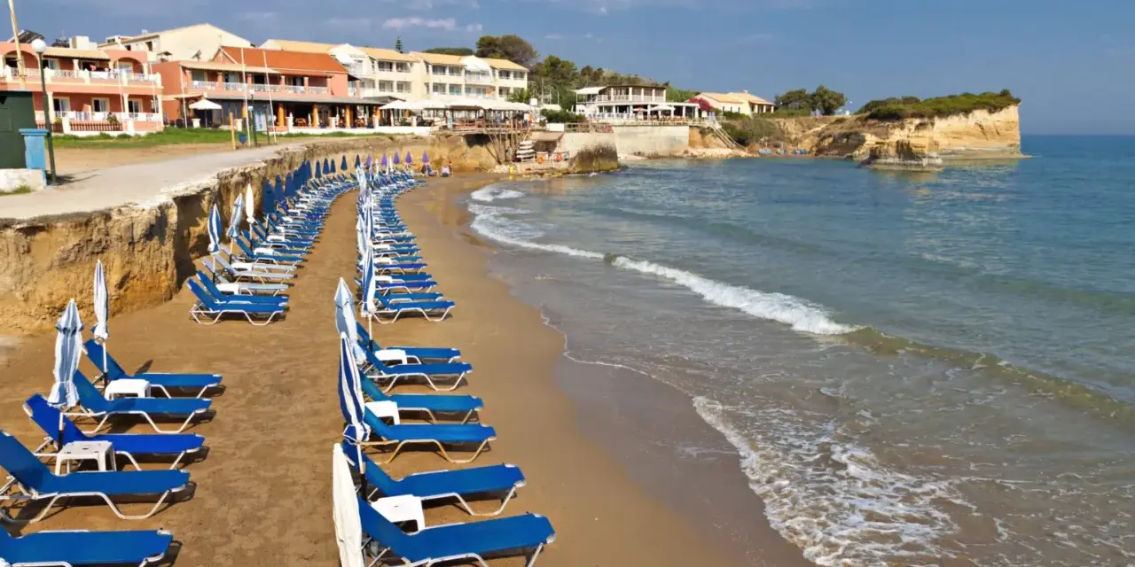 Rzędy leżaków i parasoli na piaszczystej plaży, z hotelami w tle. Idealne miejsce, by odpocząć i zastanowić się, gdzie na Korfu wybrać się na wakacje.