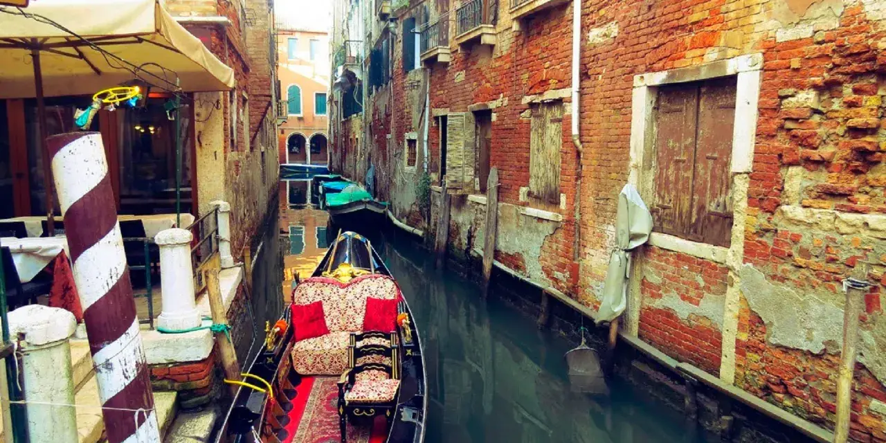 Gondola z czerwonymi poduszkami i dywanem w wąskim kanale Wenecji, otoczonym starymi ceglanymi budynkami.