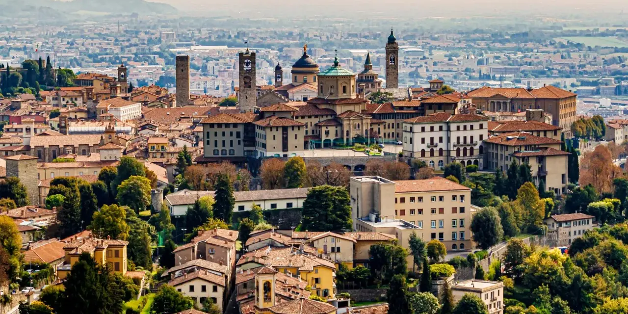 Widok na **bergamo stare miasto** z wieżami i kopułami, otoczone zielenią i innymi budynkami.