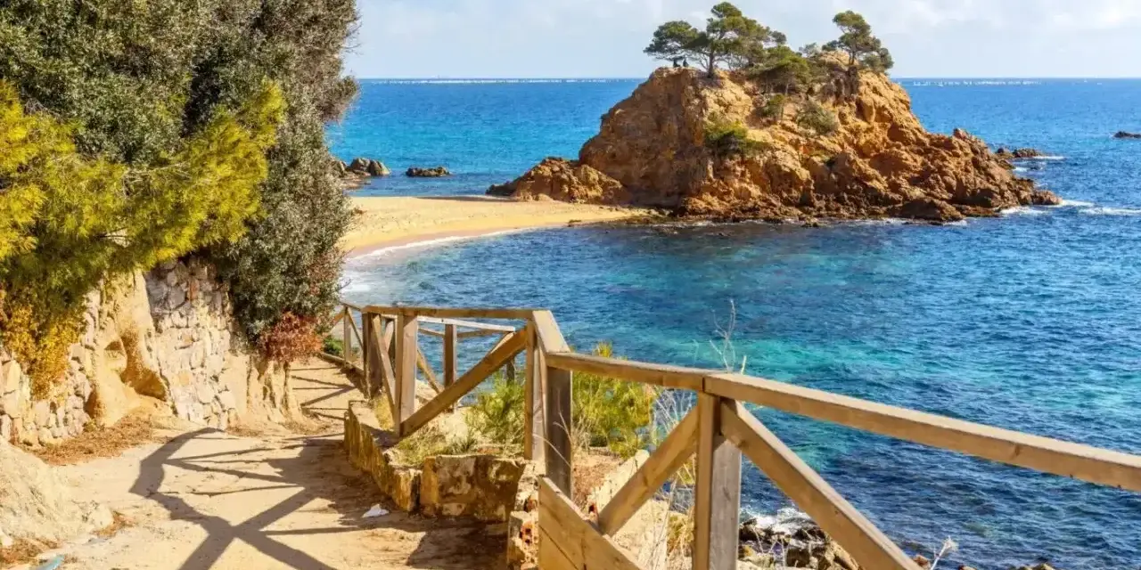 Ścieżka z drewnianą balustradą prowadzi do urokliwej zatoczki z piaszczystą plażą i skalistą wysepką. To idealne miejsce na odkrywanie uroków Costa Brava.