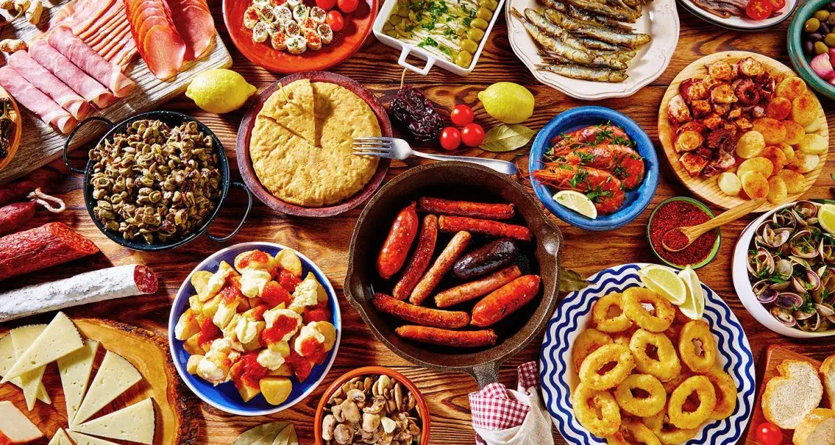 Tapas co to? Obficie zastawiony stół pełen hiszpańskich przekąsek: kiełbaski, krewetki, kalmary, owoce morza, sery, wędliny i tortilla.