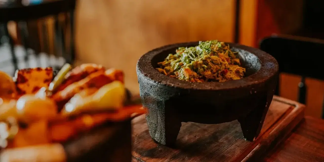Świeże guacamole w kamiennym moździerzu, obok talerza z innymi najpopularniejszymi daniami meksykańskimi.