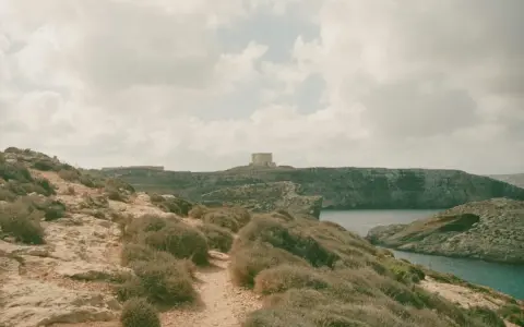 Malta czy warto? Kamienista ścieżka prowadzi przez skaliste wybrzeże z niską roślinnością do zatoki. W oddali widać wieżę obronną.