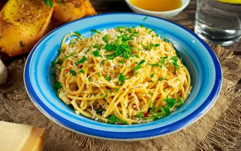 Pyszne spaghetti aglio olio posypane parmezanem i natką pietruszki, podane w niebieskiej misce. Obok grzanki czosnkowe i kawałek sera.
