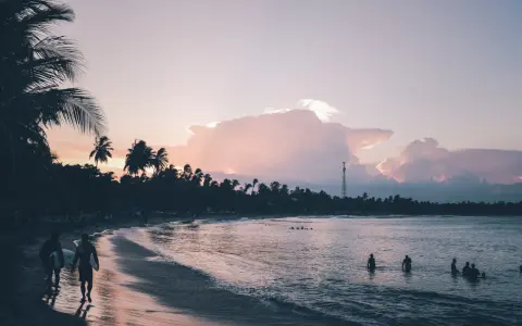 Zachód słońca na Sri Lance we wrześniu. Surferzy idą plażą, w oceanie kąpią się ludzie. Palmy i chmury w pastelowych barwach.