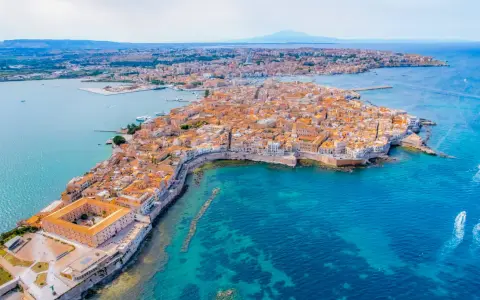 Sycylia w kwietniu: malownicza wyspa Ortigia z kamiennymi budynkami i turkusowym morzem, w oddali widać wulkan.