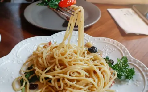 Widok z bliska na talerz z makaronem aglio olio, z widelcem unoszącym porcję spaghetti z bazylią i pomidorkiem.