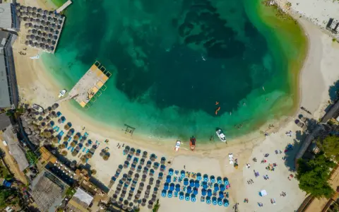 Widok z lotu ptaka na zatokę z turkusową wodą, plażą z leżakami i parasolami oraz łodziami.