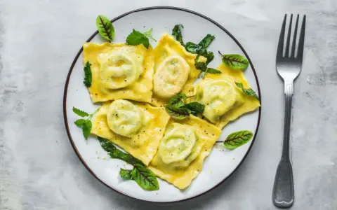 Pyszne ravioli z ricottą i szpinakiem, polane sosem, ozdobione świeżymi ziołami. Obok leży widelec.