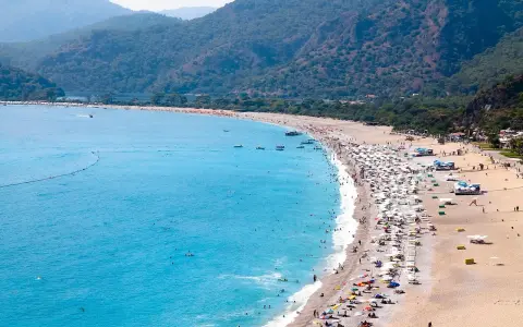 Plaża z turkusową wodą, górami w tle i tłumem ludzi odpoczywających pod parasolami.