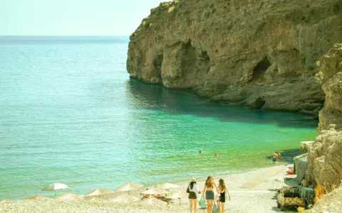 Trzy dziewczyny idące po kamienistej plaży w kierunku turkusowego morza przy skalistym klifie.
